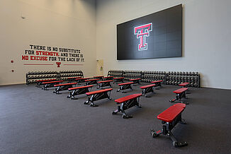 The free-weight area of the Texas Tech Football weight room, featuring a number of benches and a vast amount of dumbbells for athletes.