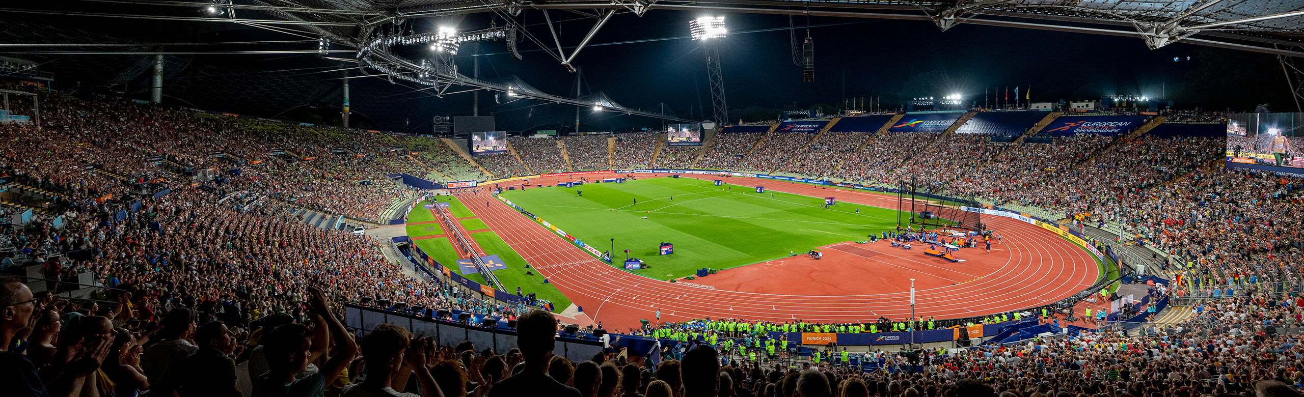 Athletics running track at Munich Olympic Stadium – red synthetic surface during the 2022 European Athletics Championships under lights. Wide-angle view of the athletics running track at Munich Olympic Stadium at night during the 2022 European Athletics Championships. The red synthetic surface is surrounded by thousands of spectators. Fully illuminated, the stadium hosts elite European track and field competition.