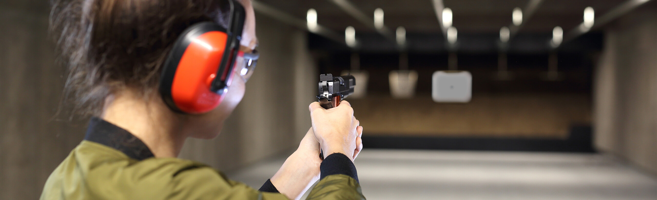 A person wearing hearing protection shoots at a target with a pistol in an indoor shooting range with a safe floor covering.
